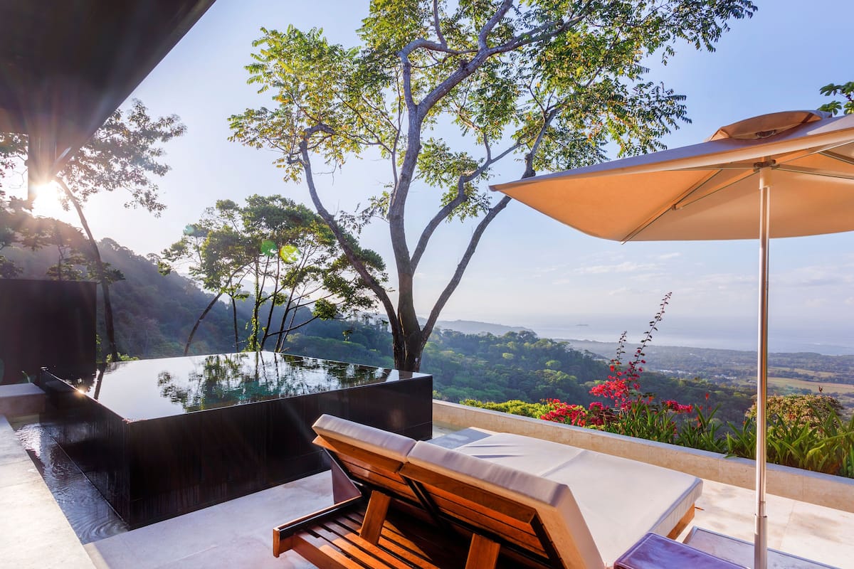 Un área lounge moderna al aire libre con una mesa de vidrio, sillones de madera y un parasol grande.