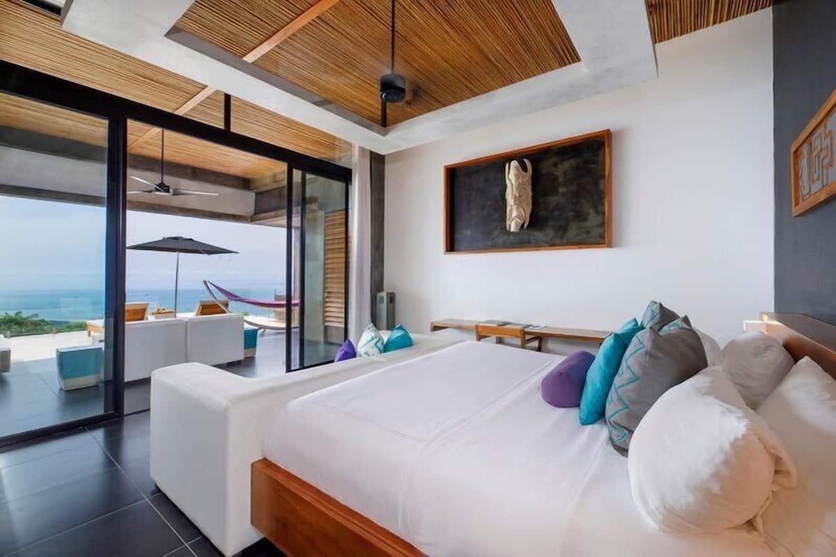 Un dormitorio moderno con una cama grande, vistas al océano y un balcón con mobiliario exterior.