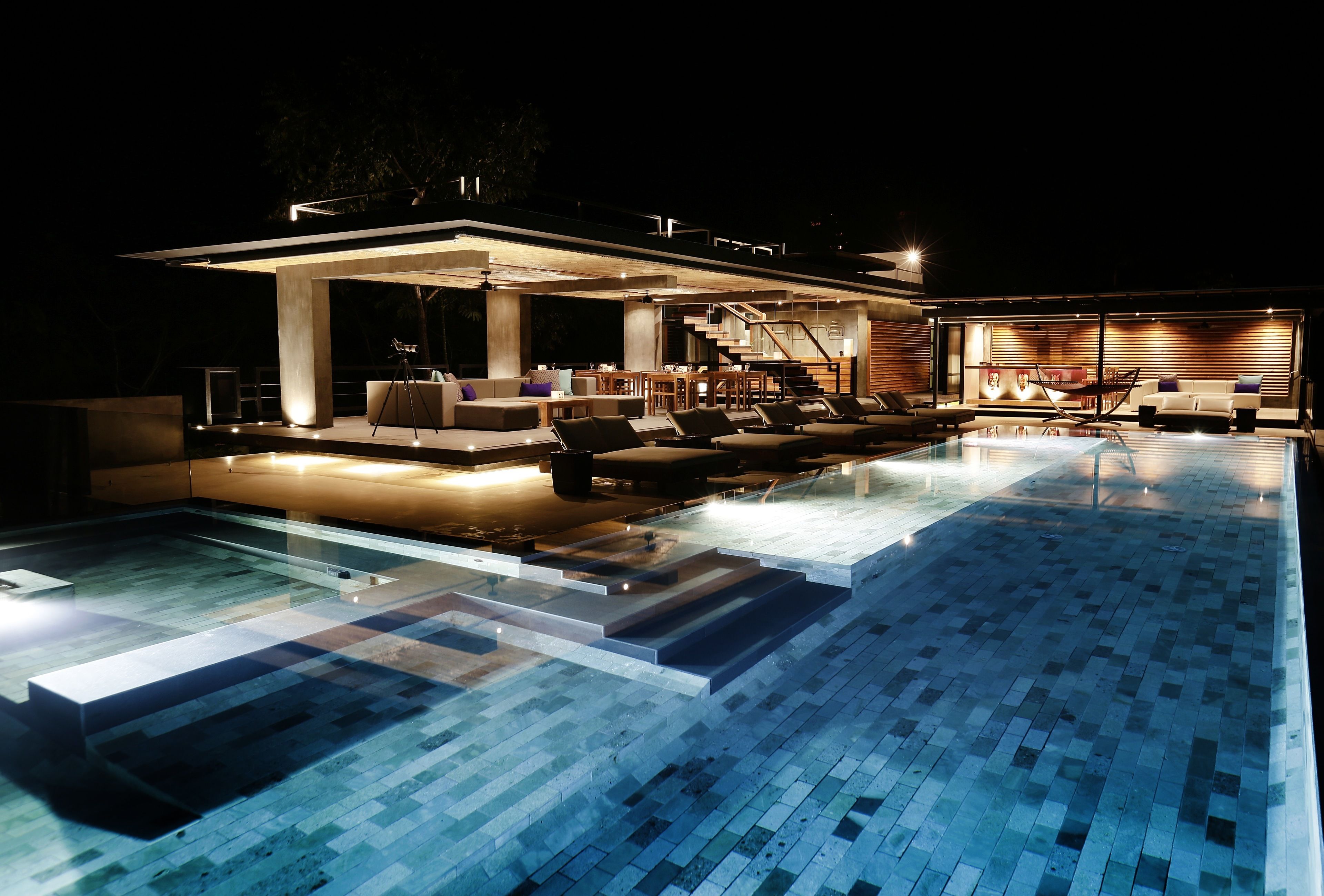 A modern outdoor pool area with a covered seating area, a staircase, and a lounge with sofas and chairs.