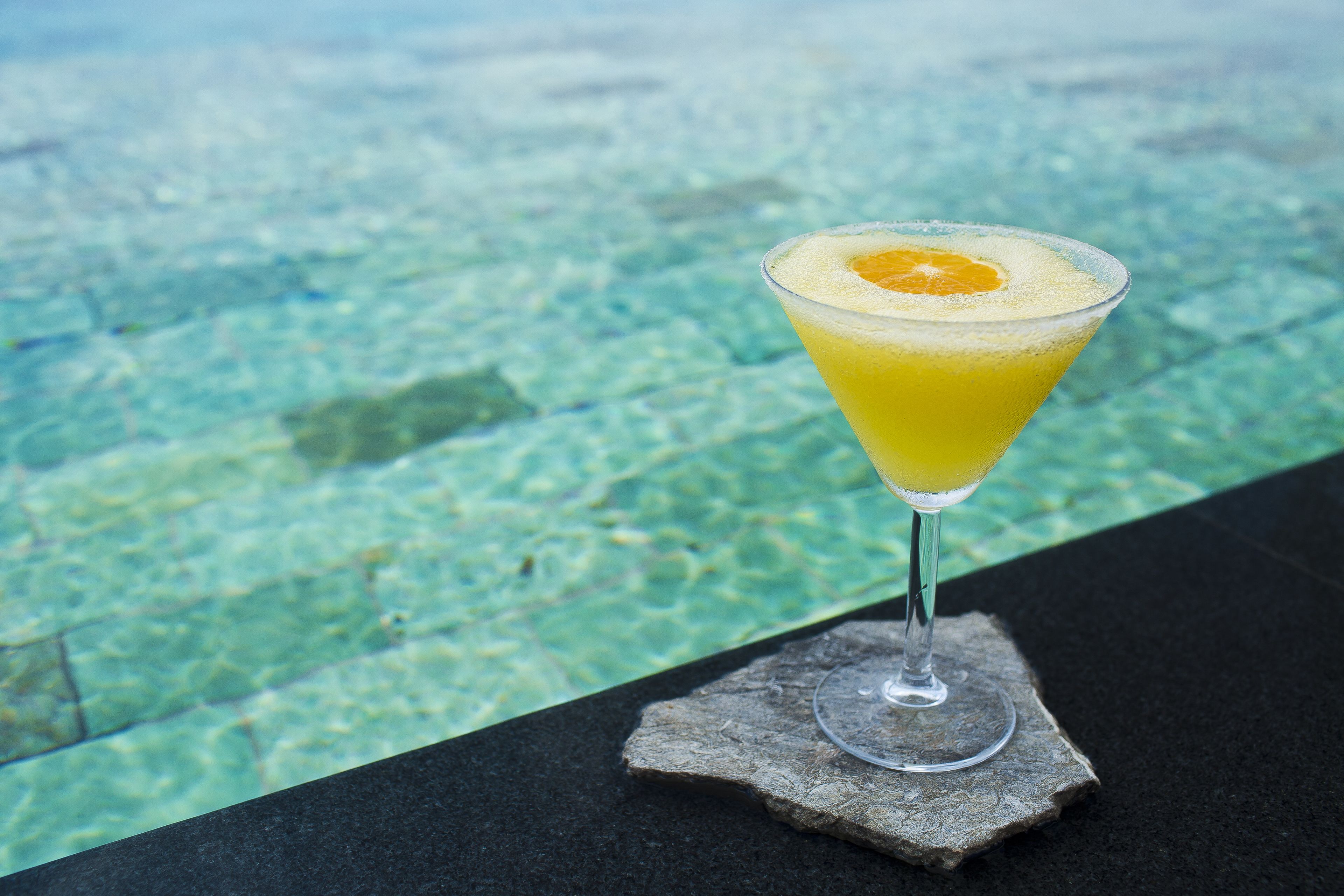 A cocktail glass with an orange garnish on a stone coaster by a pool.