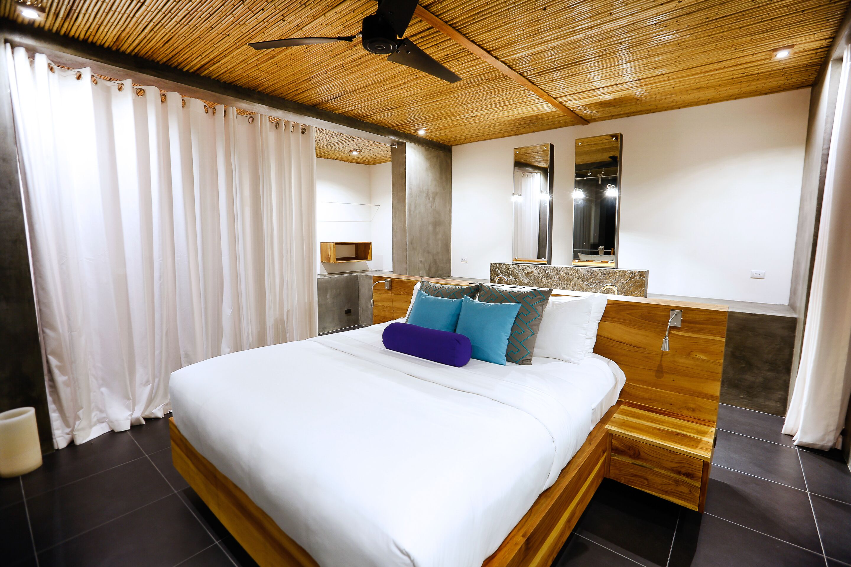 Ein modernes Schlafzimmer mit einem hölzernen Kopfteil, einem Bett mit weißer Bettwäsche, einem Deckenventilator und einem großen Spiegel über dem Bett.