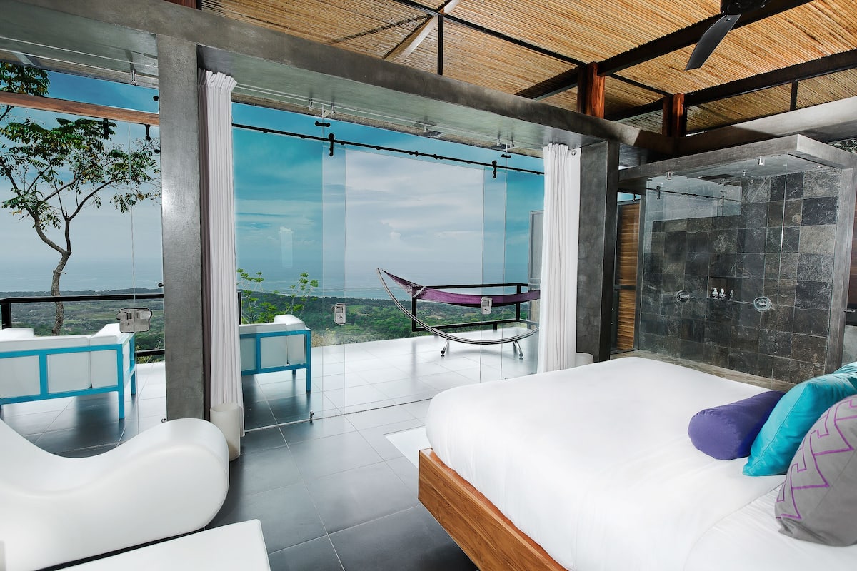 Un dormitorio moderno con una cama grande, cabina de ducha de vidrio y balcón con vistas al paisaje.