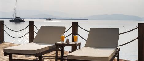 Two lounge chairs on a wooden deck with a view of the sea and a sailboat in the distance.