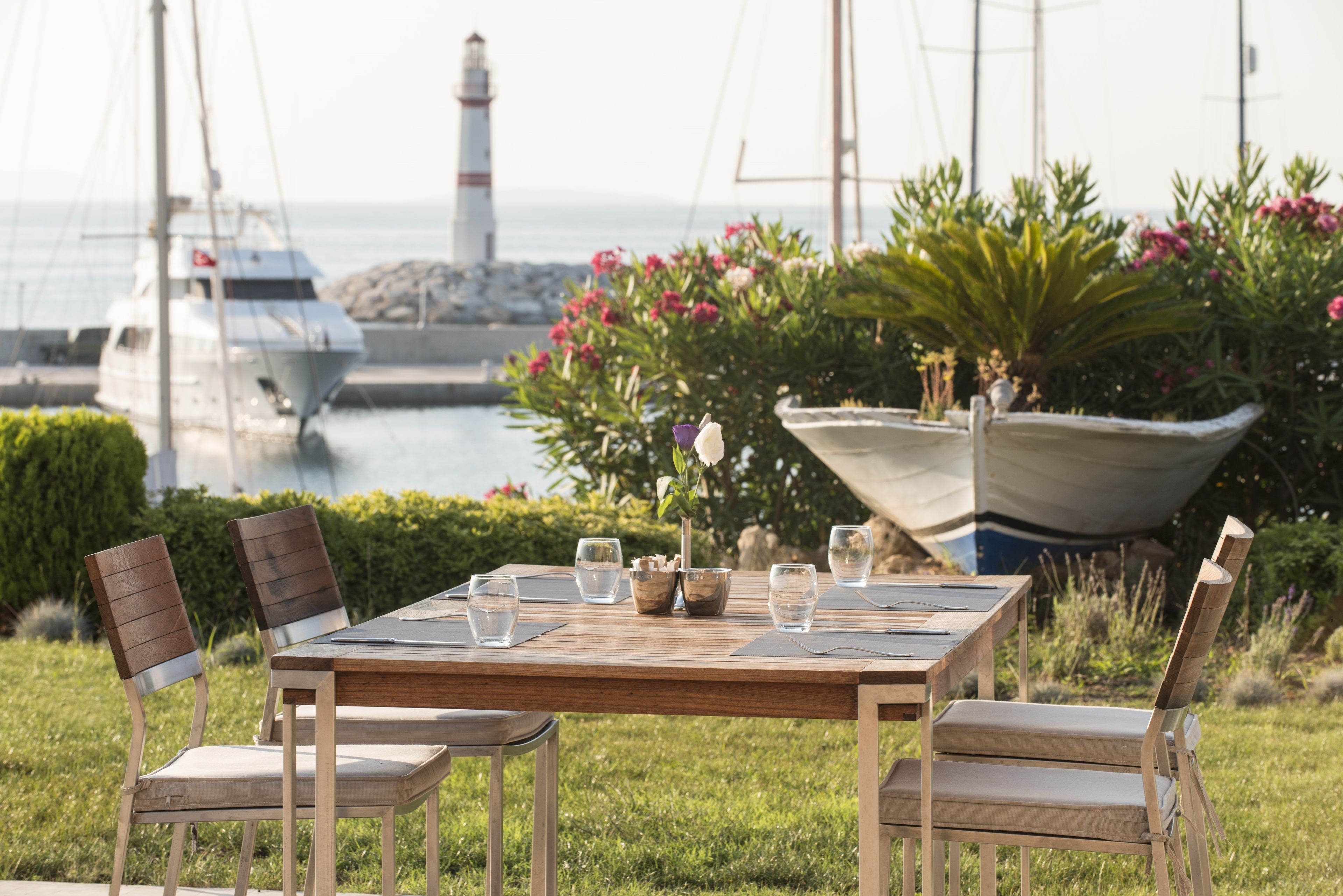 An outdoor dining area with a table and chairs, a flower vase, and a view of boats and a lighthouse.