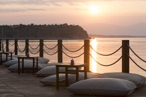 A wooden deck with tables and pillows, a rope barrier, and a sunset over water. - Yacht Club (Didim)