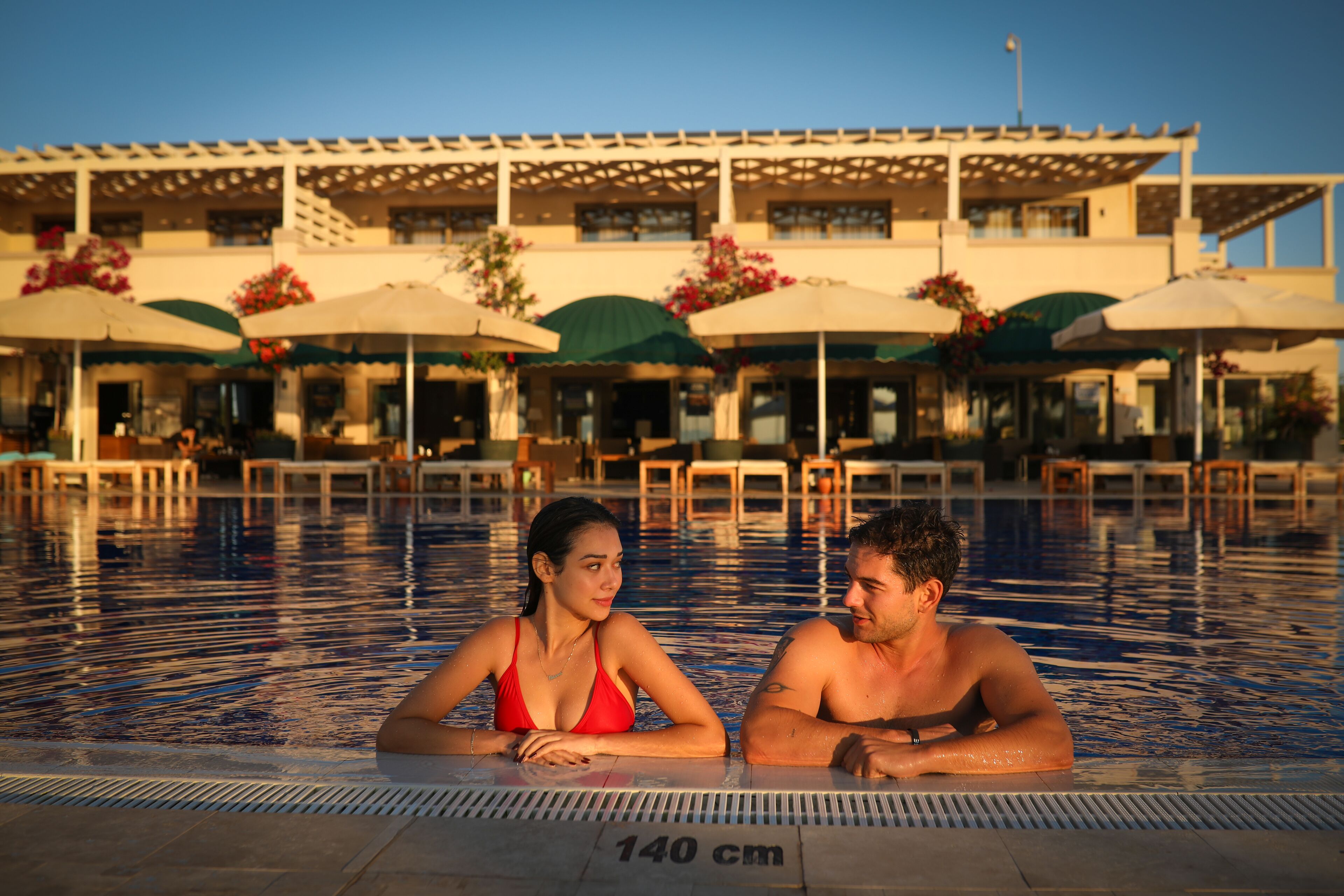 A swimming pool with a 140 cm depth marker, surrounded by outdoor seating with umbrellas and a building in the background.