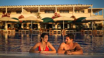 A swimming pool with a 140 cm depth marker, surrounded by outdoor seating with umbrellas and a building in the background.