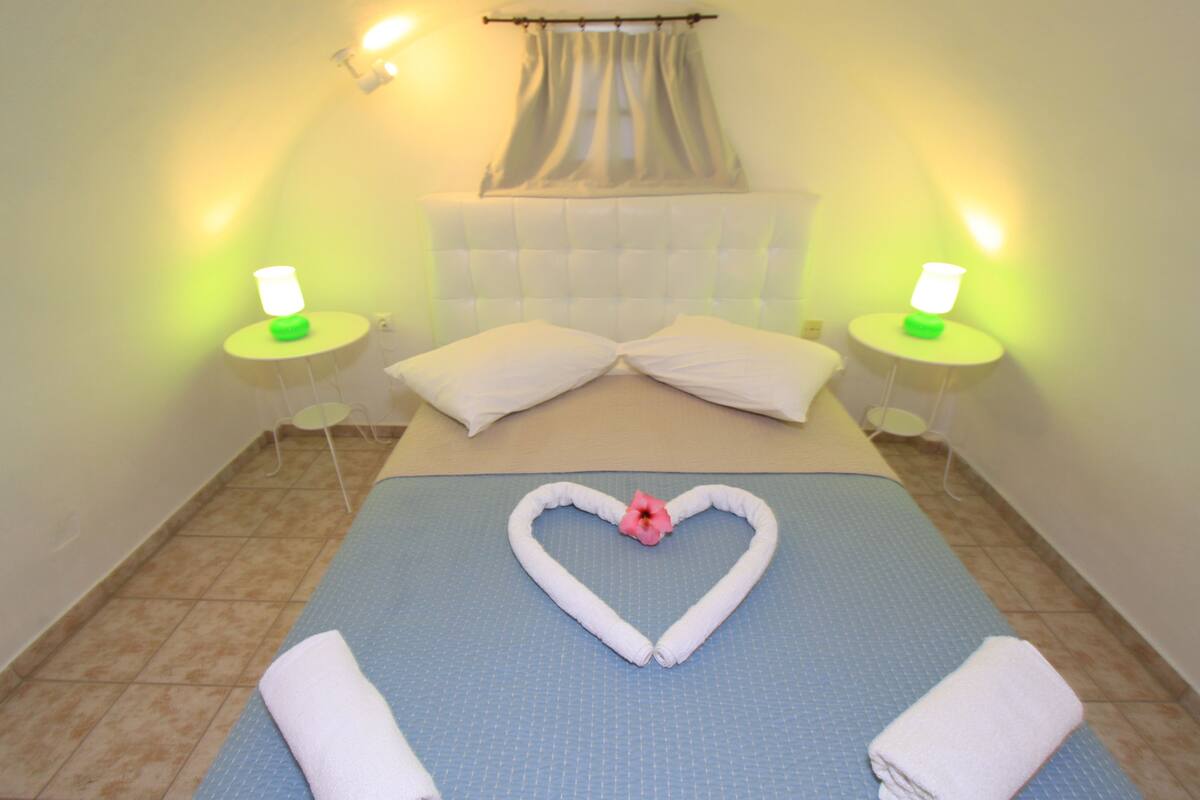 A hotel room with a bed, two bedside tables with lamps, a heart-shaped towel decoration, and a window with curtains.