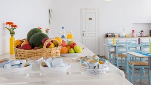 Un bufé de desayuno con frutas, pasteles y bebidas sobre una mesa blanca en una habitación luminosa con sillas azules y paredes blancas.