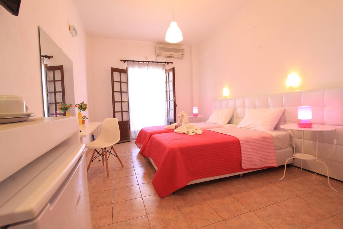 A hotel room with two beds, a desk, a chair, and a small table with a lamp.
