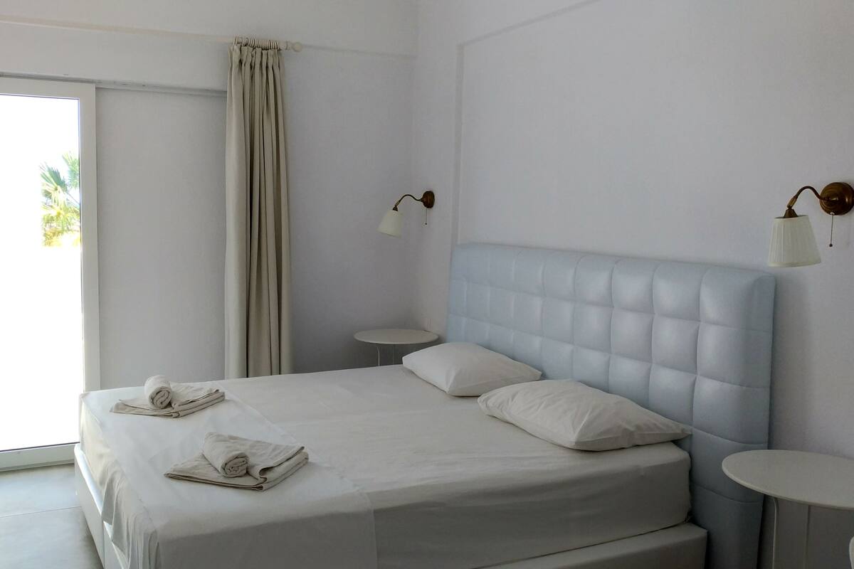 A neatly made bed with two pillows and two folded towels, a bedside table, a round table, and a window with curtains.