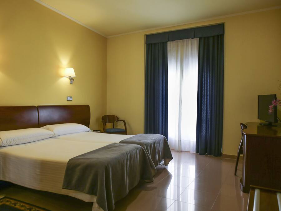 Habitación de hotel con dos camas, un escritorio, una silla y un ventanal con cortinas.