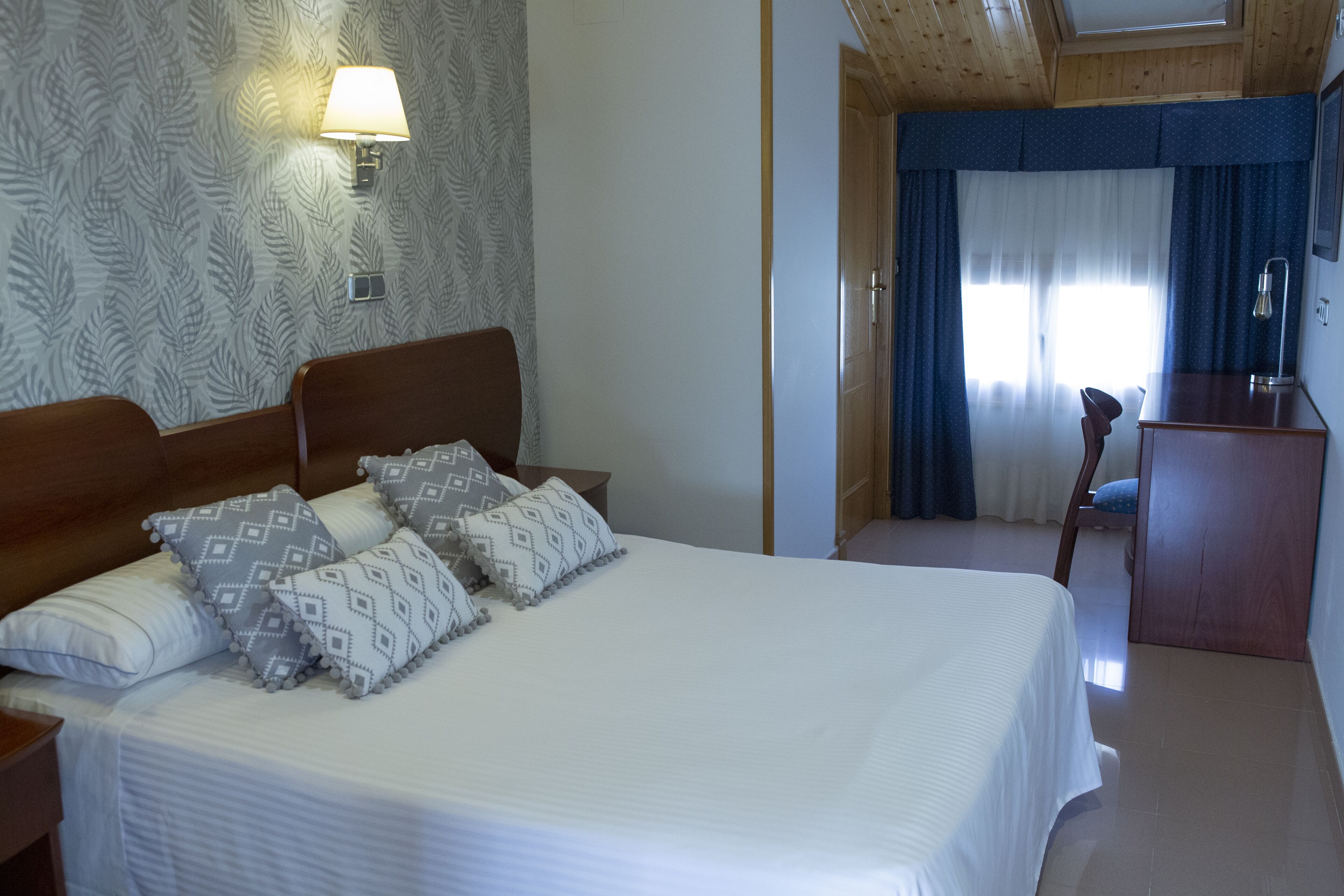 Una cama bien hecha con almohadones estampados, una lámpara de noche y un cabecero de madera.