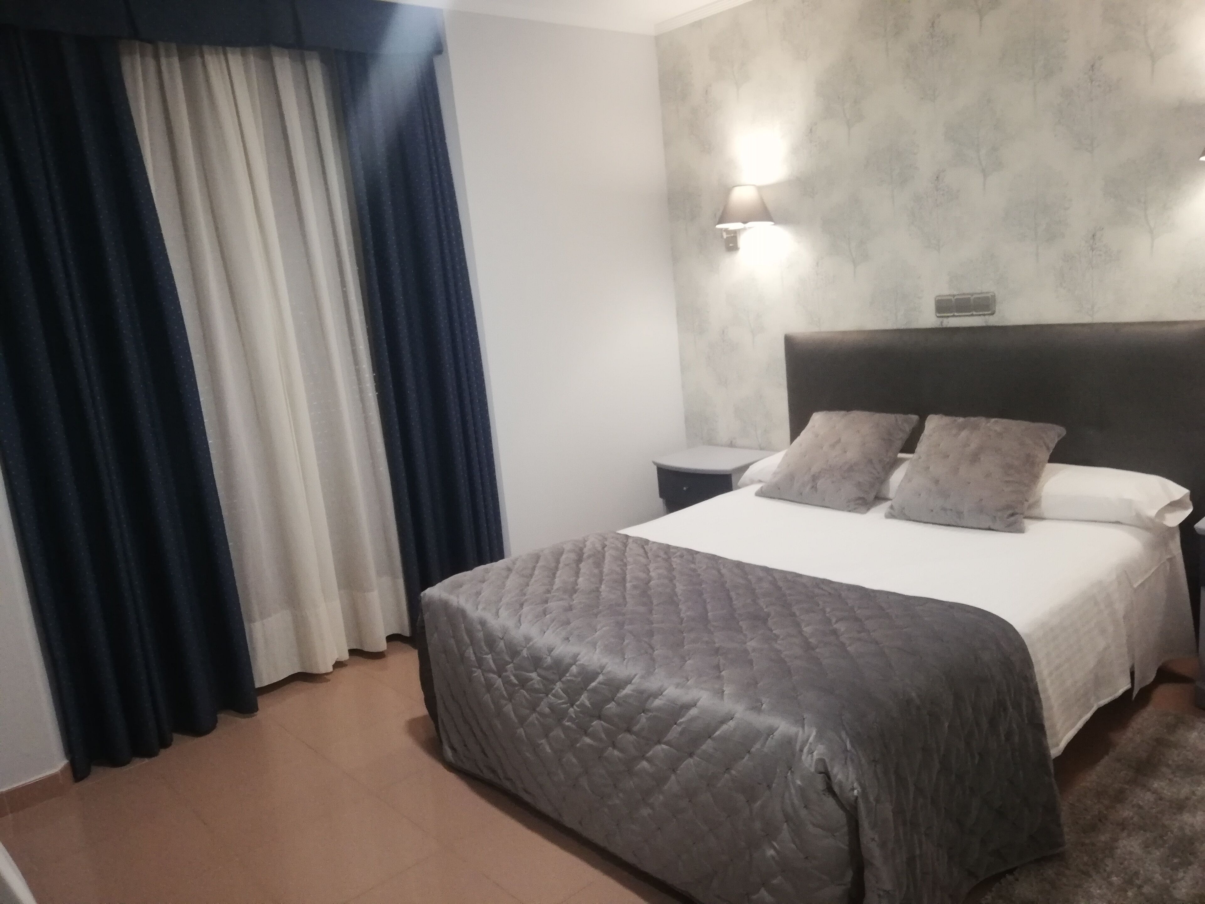 Une chambre d’hôtel avec un lit, une table de chevet, une lampe fixée au mur et une fenêtre avec des rideaux.