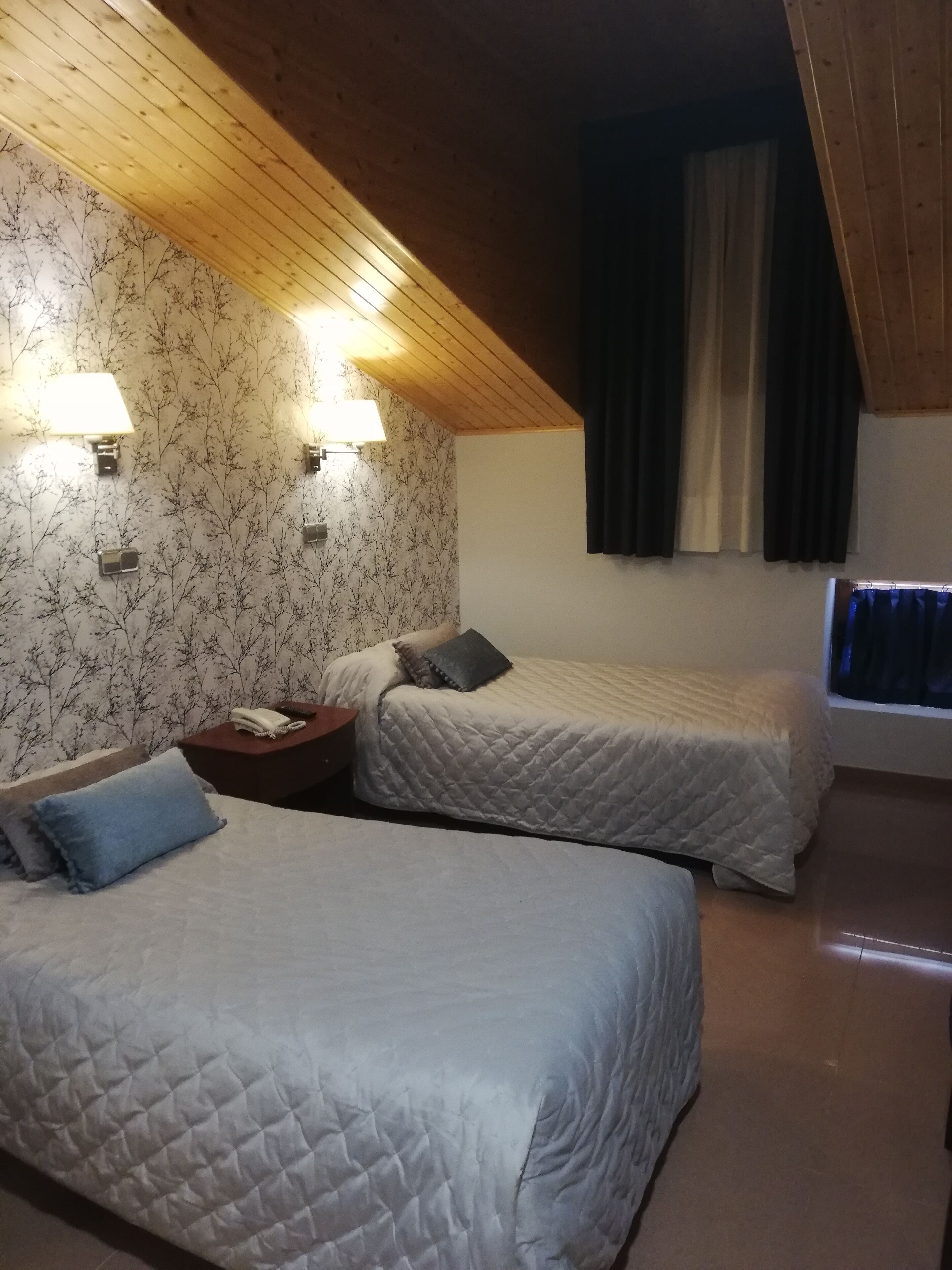 Ein Zimmer mit zwei Betten, Holzboden, Wandlampen, einem kleinen Nachttisch mit Telefon und einem Fenster mit Vorhängen.