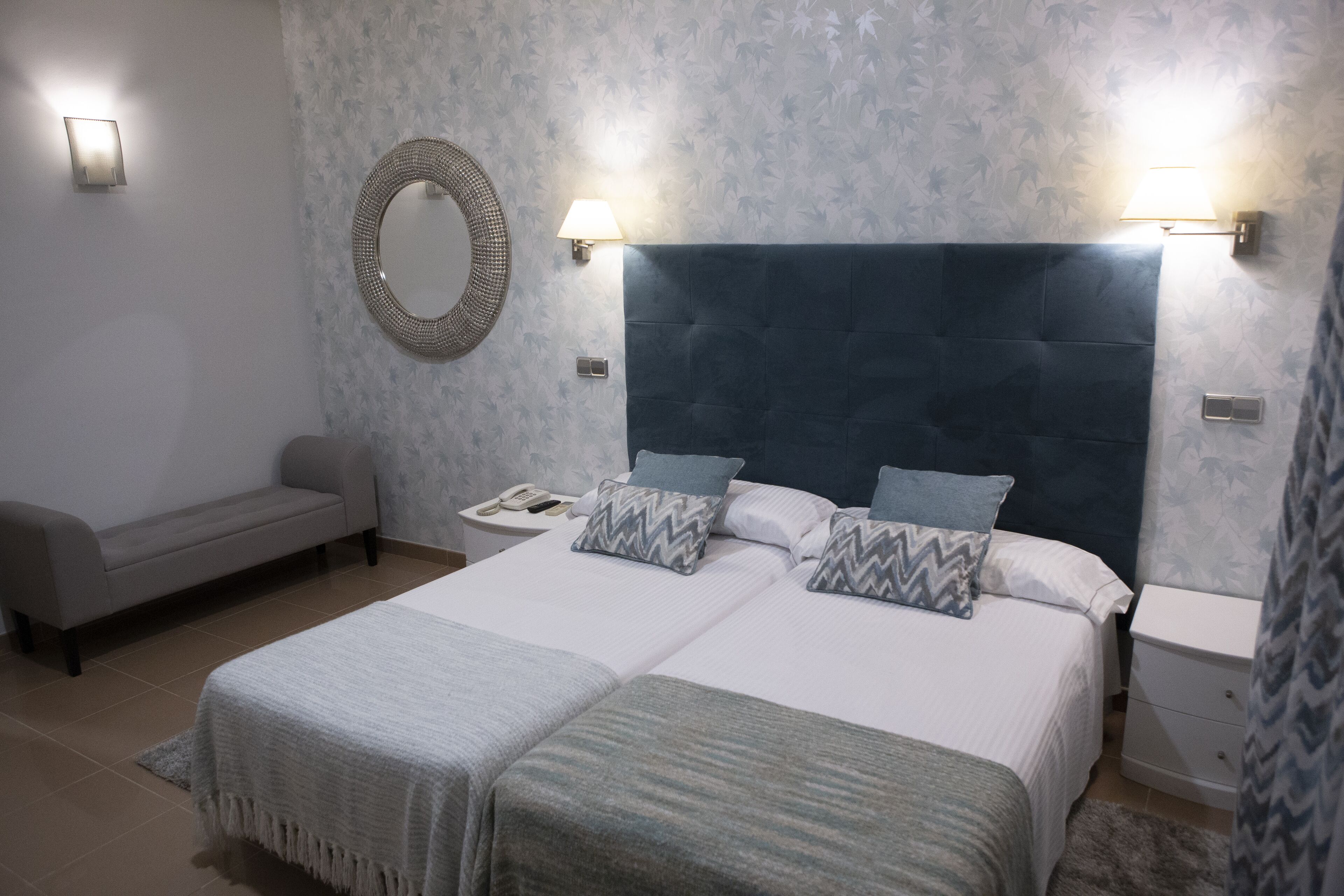 Una cama doble con cabecera oscura, dos mesitas de noche, una mesita auxiliar, un espejo fijado a la pared y un sofá gris.