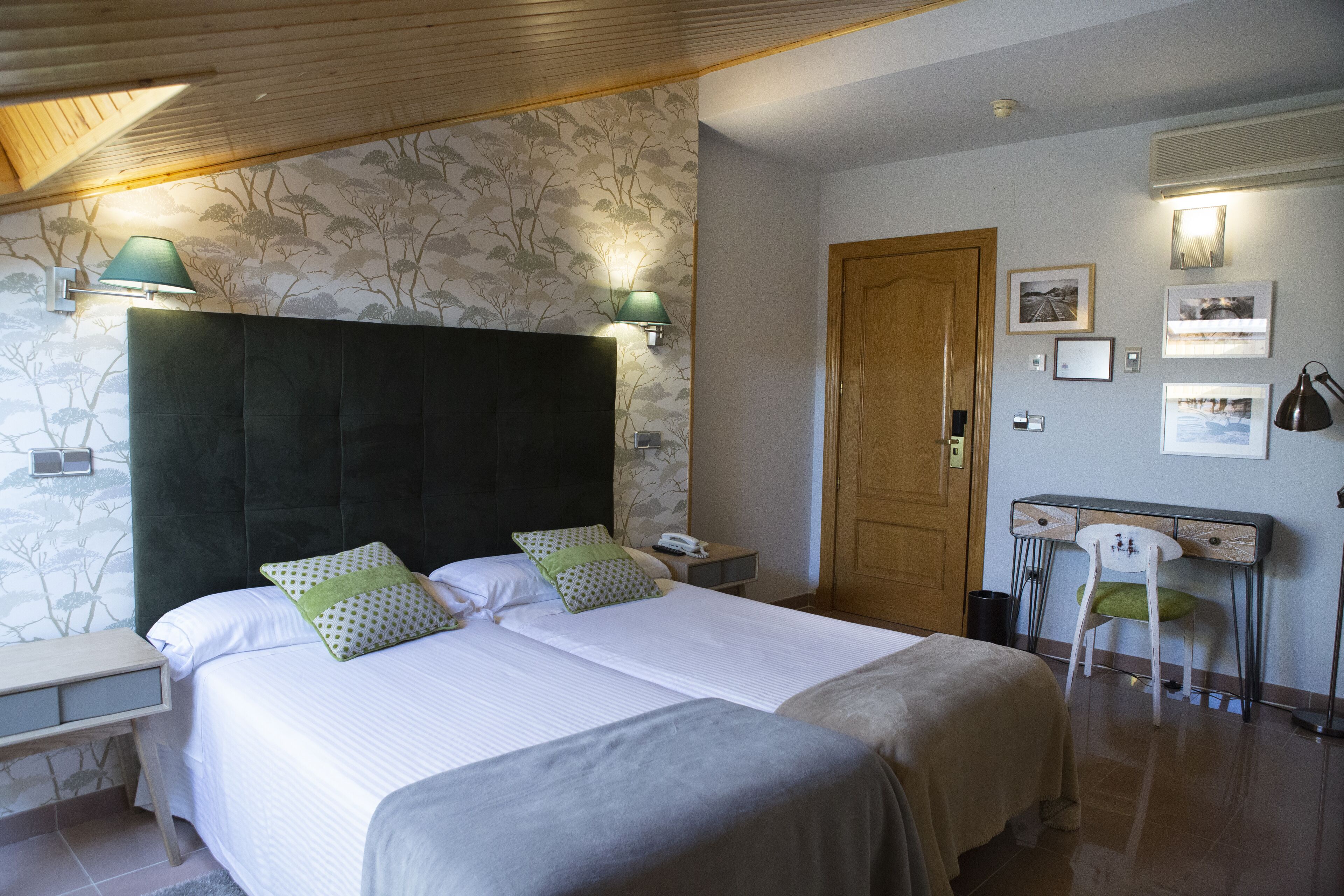 A double bed with a dark headboard, two bedside tables, a nightstand, a desk with a chair, and a wooden door.