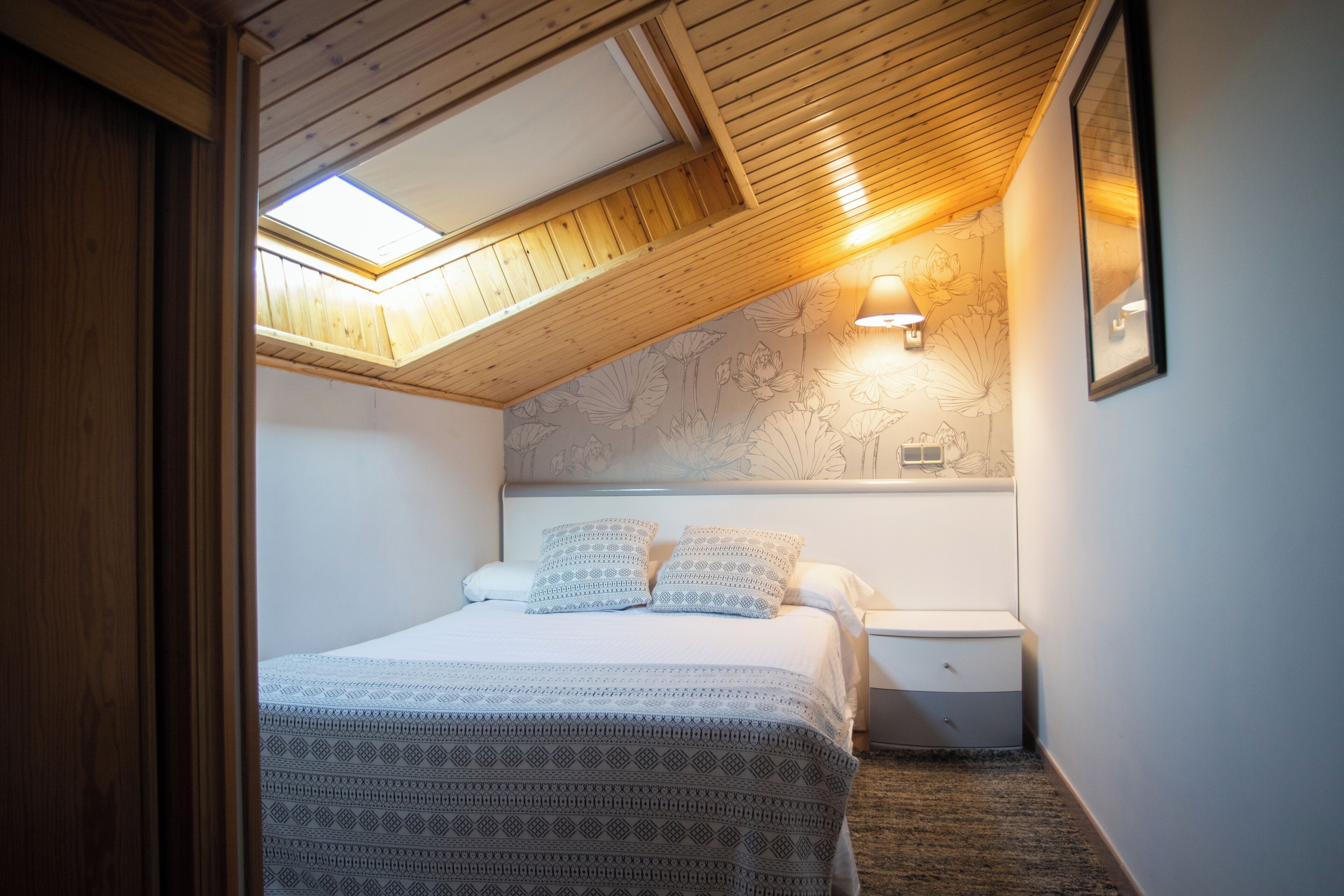 A bedroom with a sloped ceiling, a skylight, a bed with patterned bedding, a nightstand, and a wall-mounted light fixture.
