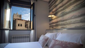 A hotel room with a large bed, a window with a view of a historic building, a wall-mounted lamp, and a radiator. - Hotel Las Moradas (Ávila)