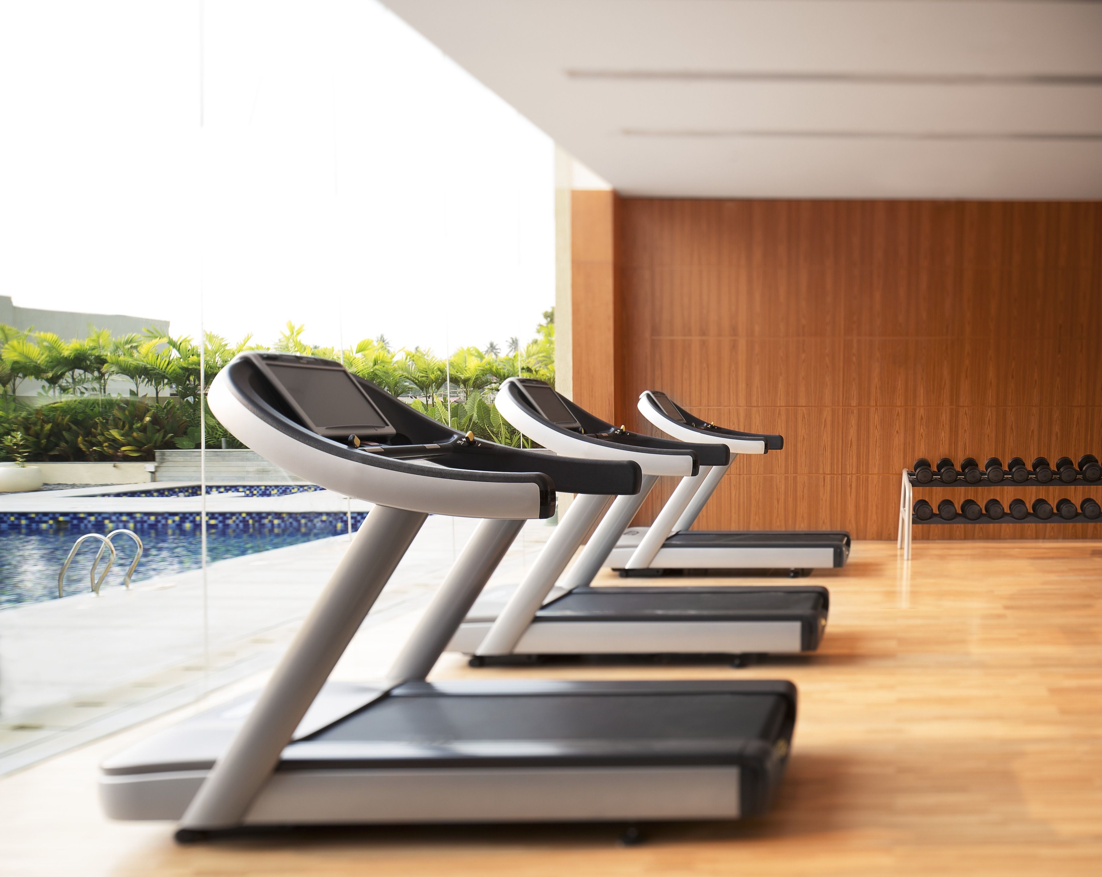 Une rangée de tapis de course dans une salle de sport, avec un plancher en bois et une vue sur une piscine.