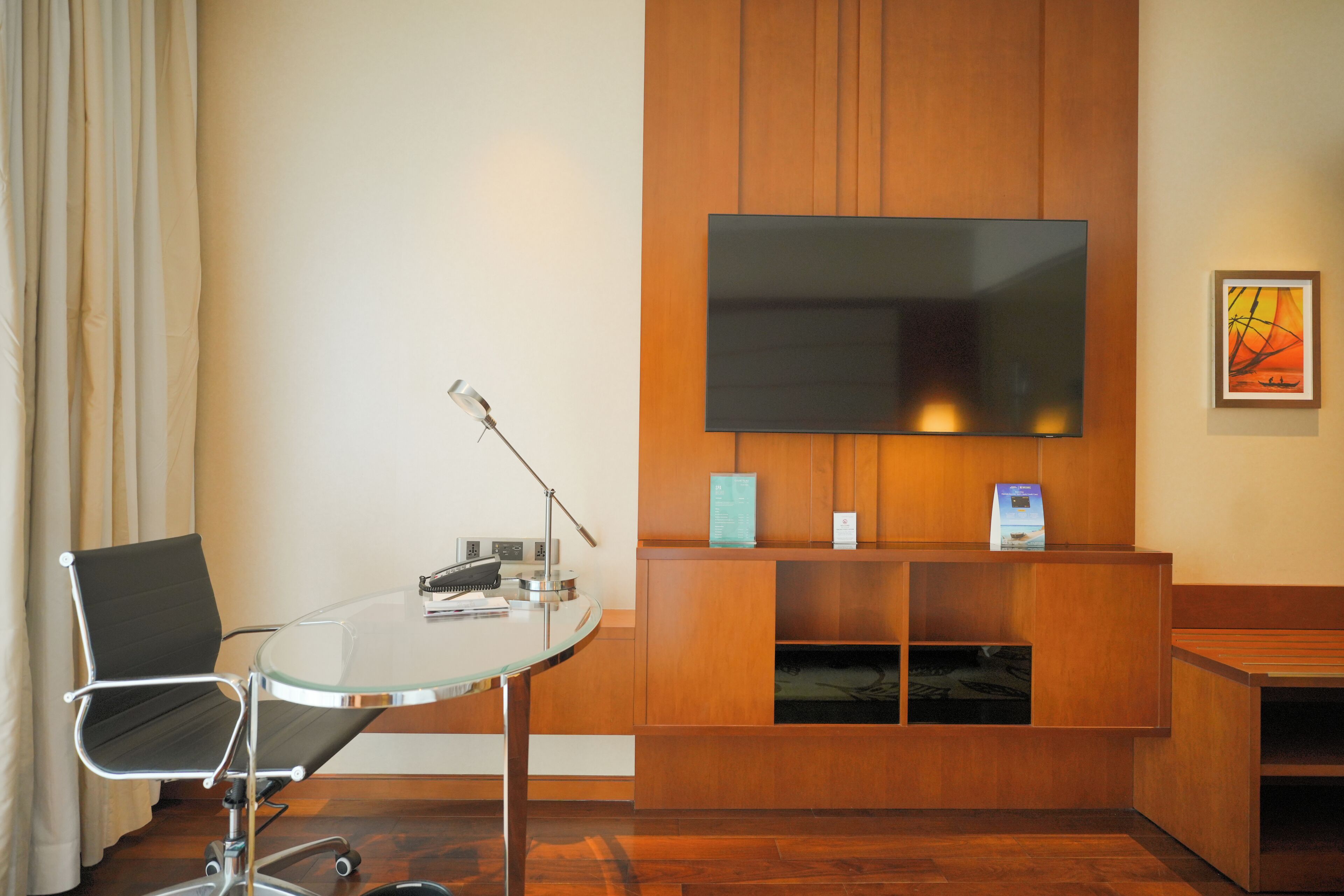 A modern hotel room with a glass table, a black office chair, a flat-screen TV mounted on a wooden panel, and a framed artwork on the wall.