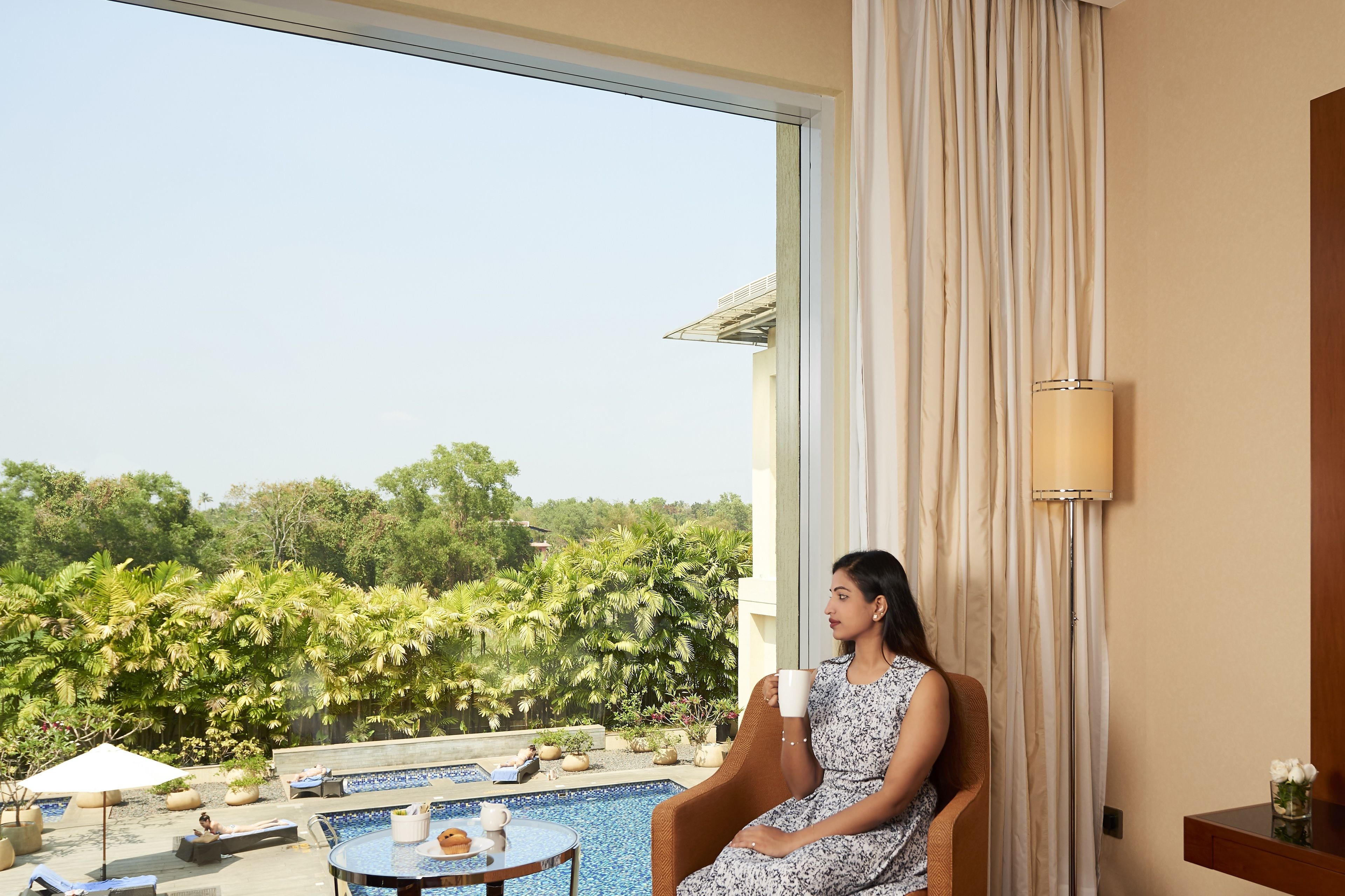 A room with a large window, a view of greenery, a pool, and a person sitting by the window.