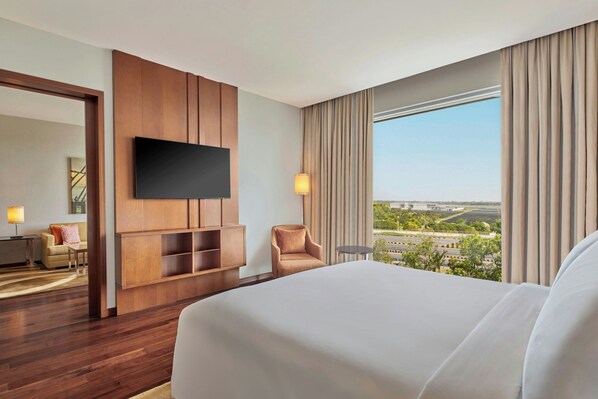 A hotel room with a large bed, a flat-screen TV mounted on a wooden panel, a small round table, a chair, and a view of the cityscape through large windows. - Courtyard by Marriott Kochi Airport (Aluva)