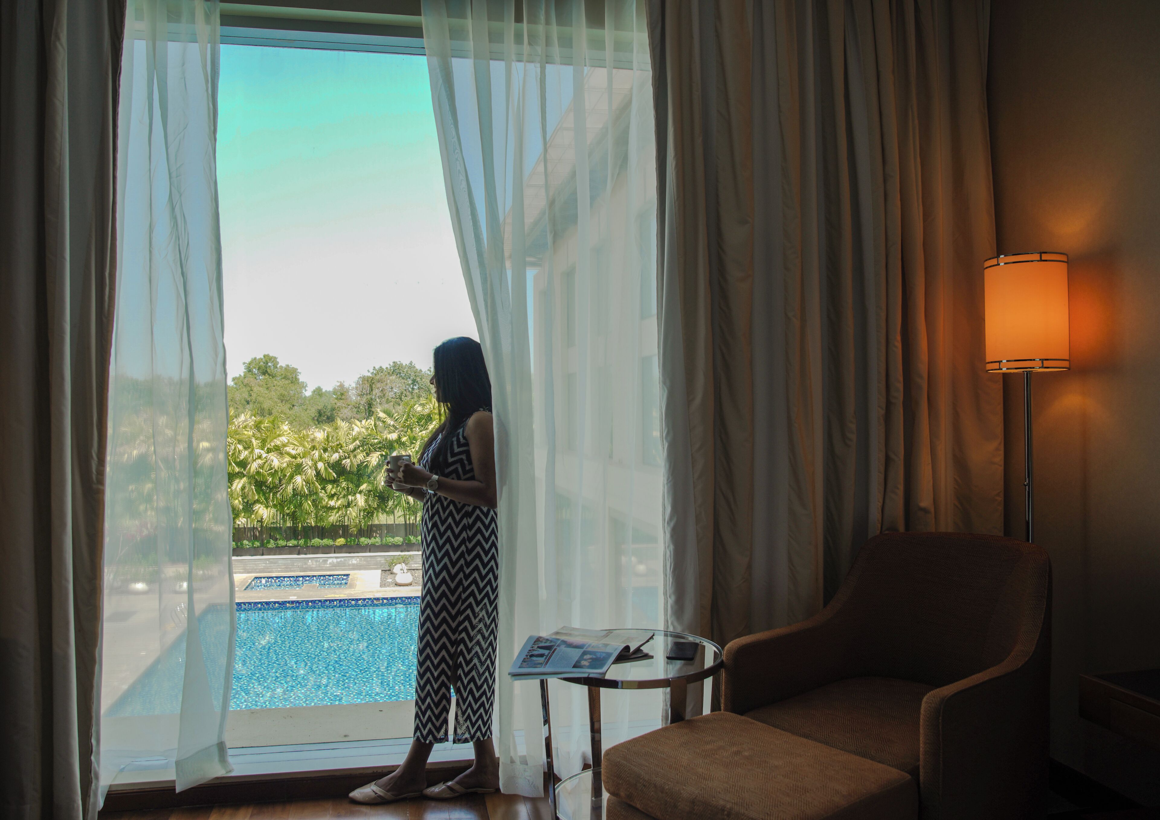 A room with a view of a swimming pool, a chair, a table with a lamp, and a person standing by a window with sheer curtains.