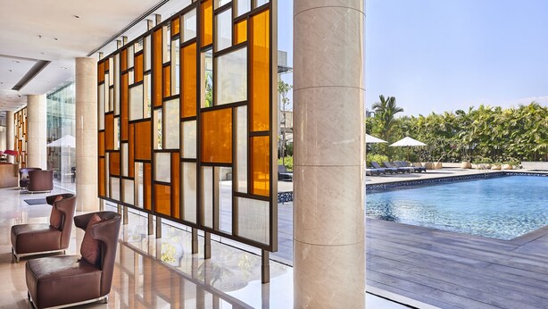 Un moderno vestíbulo de hotel con zona de piscina, destacando un amplio panel de vidrio con patrones geométricos, asientos cómodos y vistas a la piscina.