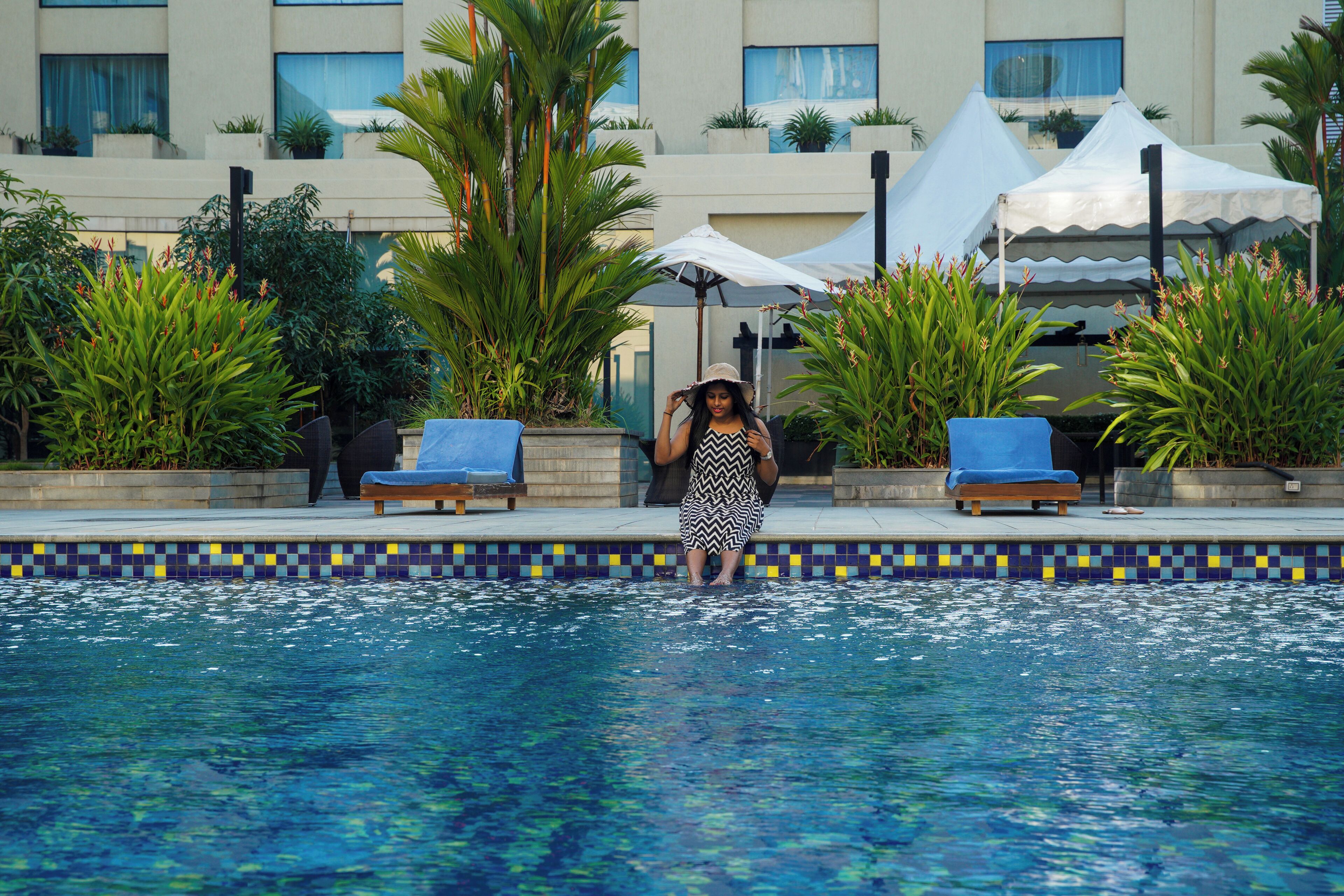 Área de piscina com espreguiçadeiras, uma pessoa perto da água e um edifício com varandas e plantas ao fundo.