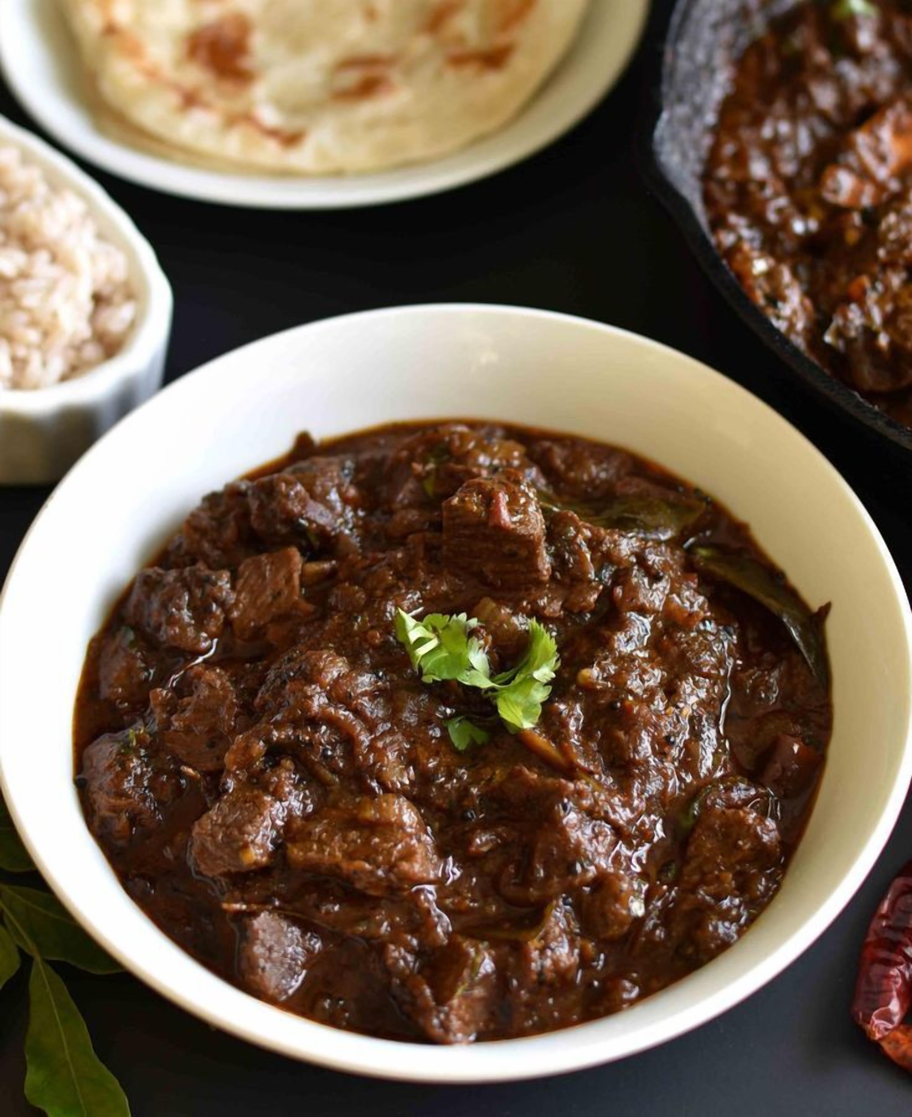 A bowl of meat-based curry with a garnish, accompanied by flatbread and a bowl of rice.