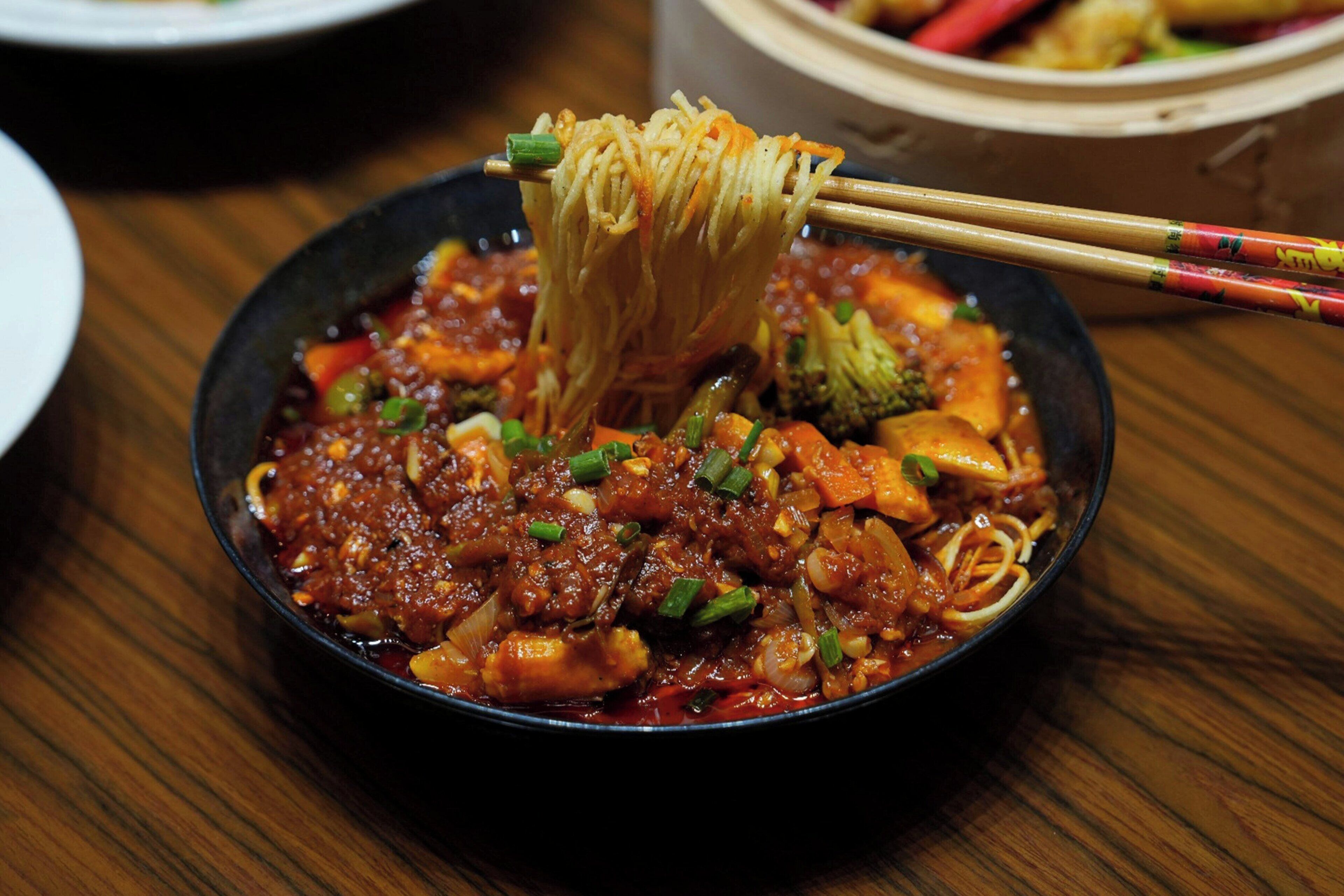 A bowl of noodles with vegetables and a sauce, being lifted with chopsticks.