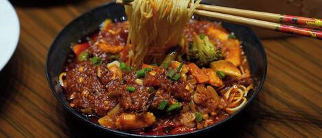 A bowl of noodles with vegetables and a sauce, being lifted with chopsticks.