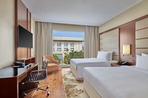 A hotel room with a desk, two beds, a TV, and a view of a building and greenery. - Courtyard by Marriott Kochi Airport (Aluva)