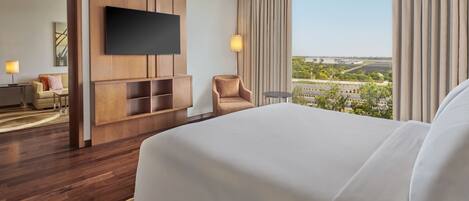 A hotel room with a large bed, a TV mounted on the wall, a seating area with a chair and a small table, and a view of the city through large windows.