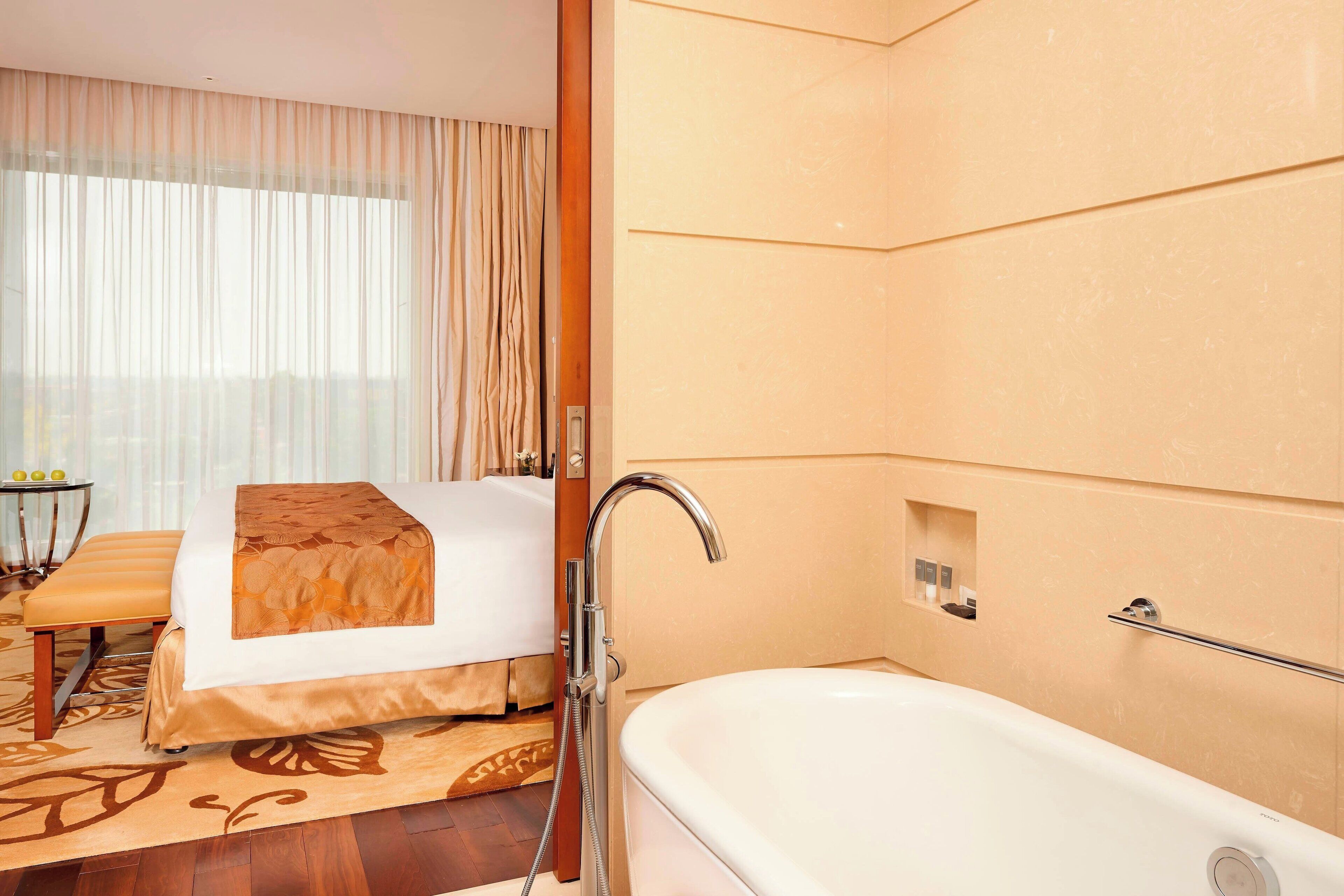 Quarto de hotel com cama, banheiro com banheira, pequena mesa com cadeira e janela com cortinas.