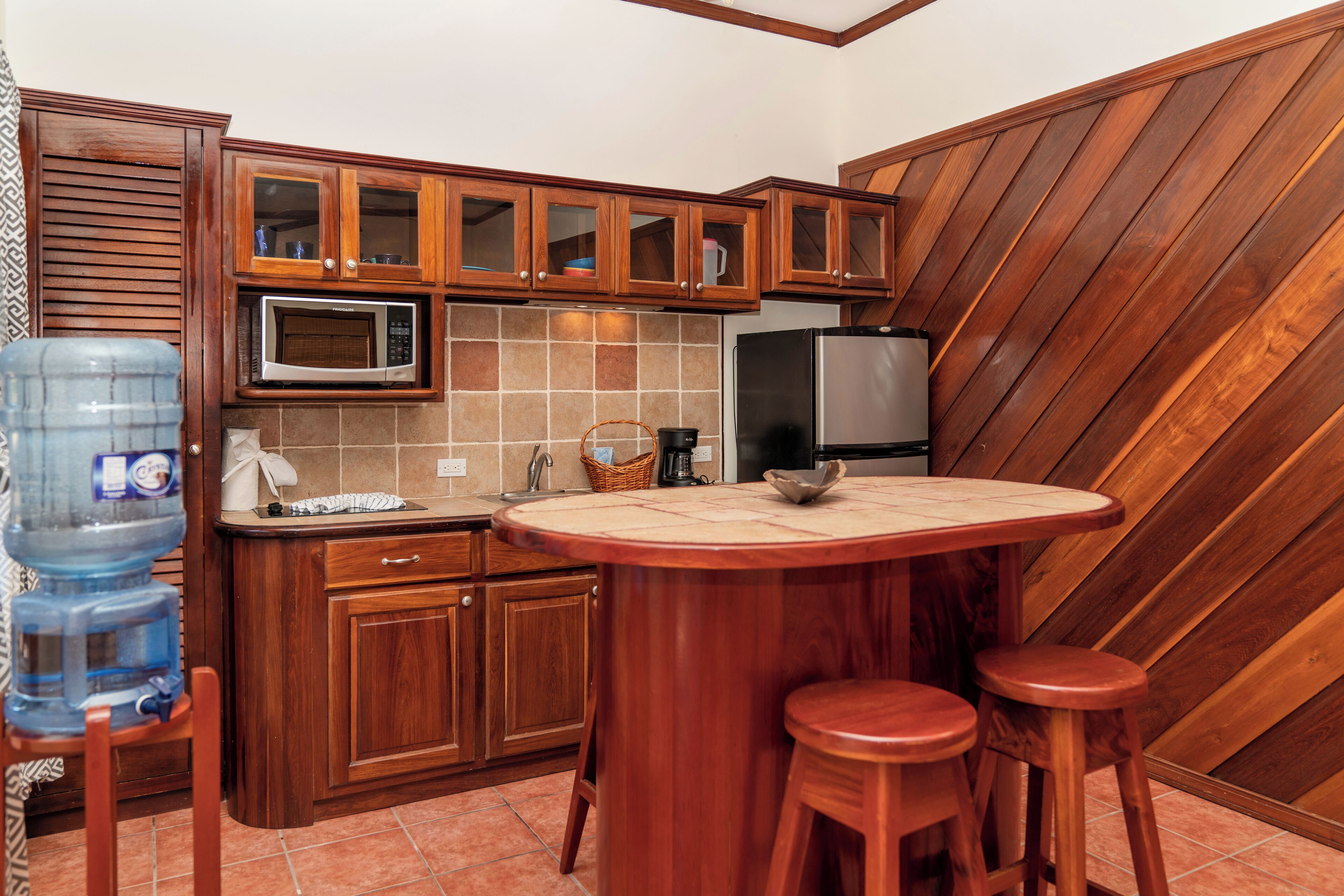 A kitchen with wooden cabinets, a microwave, a refrigerator, a water dispenser, and a round table with stools.