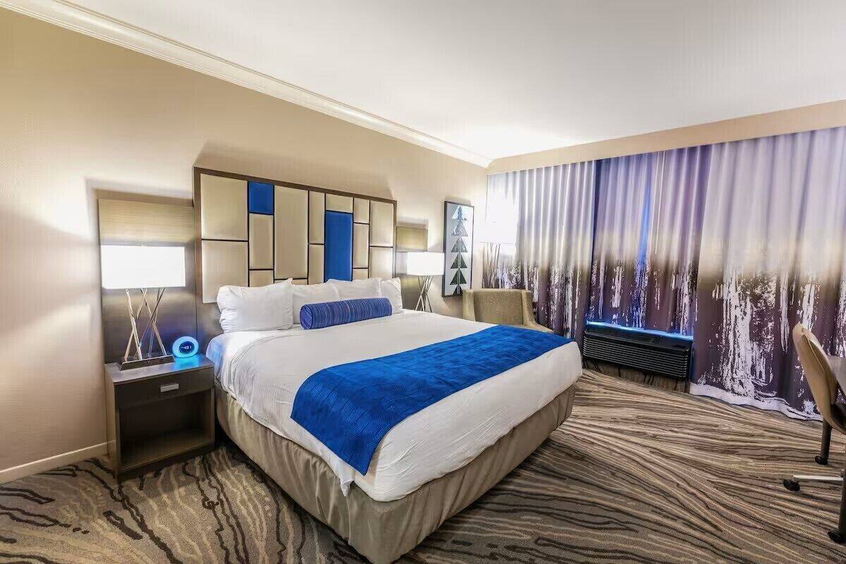 Una habitación de hotel con una cama grande, una mesita de noche, una lámpara de escritorio, una silla y vistas a la ciudad desde la ventana.