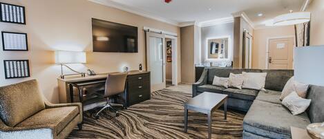 A modern hotel room with a zebra-patterned carpet, a sofa, a chair, a desk with a lamp, and a flat-screen TV mounted on the wall.