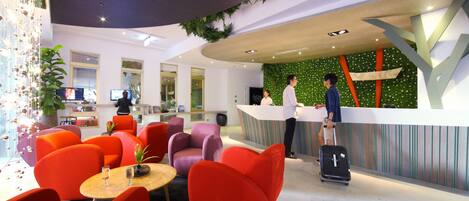 Un hall d’hôtel moderne, avec des sièges aux couleurs vives, rouge et violet, un comptoir d’accueil et un mur vert agrémenté d’un jardin vertical.