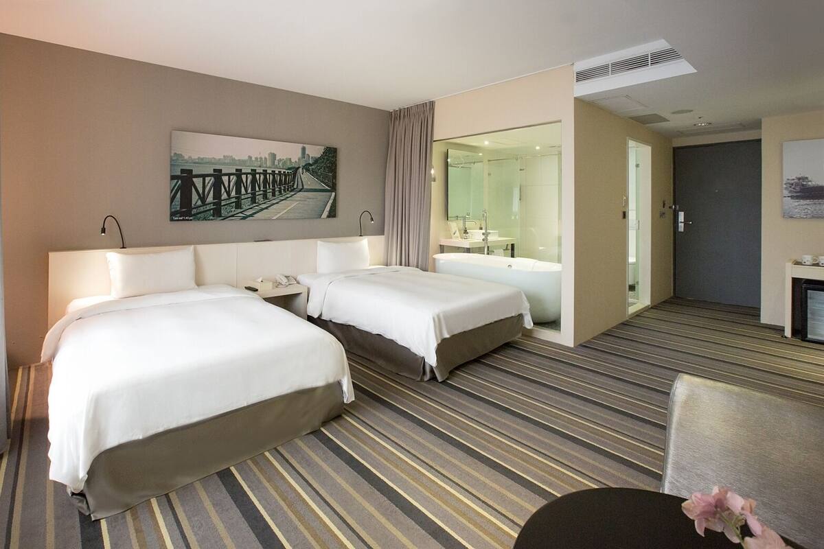 Hotelkamer met twee bedden, een groot raam met uitzicht op de stad, een badkamer met bad en een bureau met stoel.