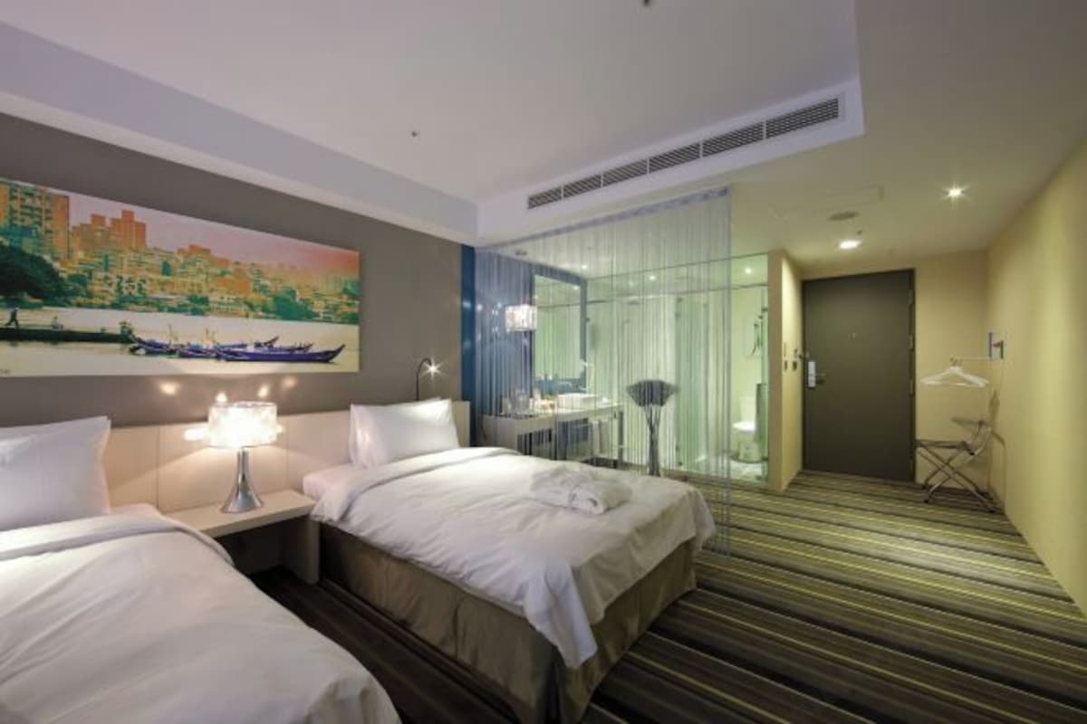 A hotel room with two beds, a painting of a cityscape, a desk with a lamp, and a view of a city through a glass door.