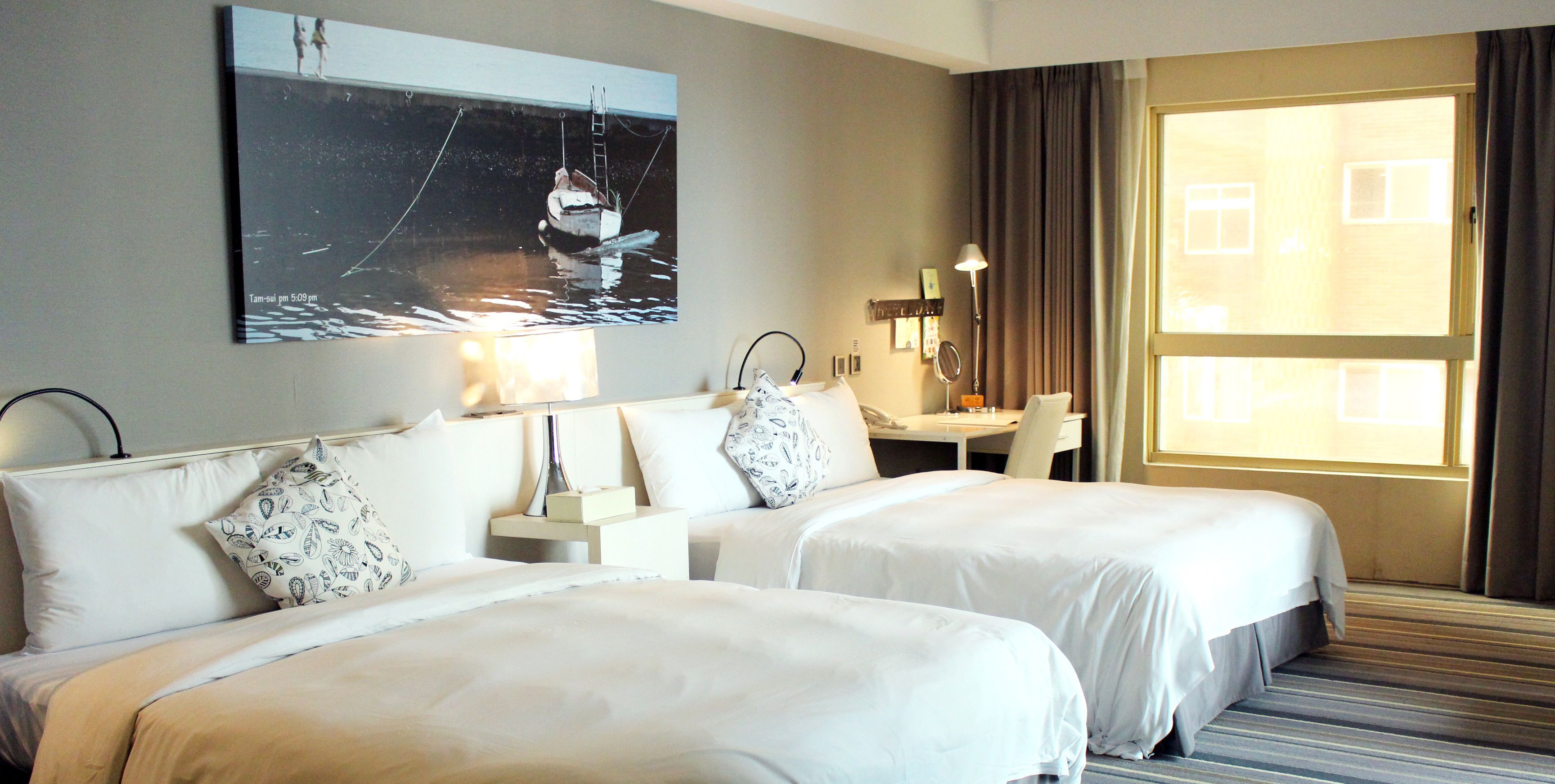 Ett hotellrum med två sängar, ett skrivbord, en stol, en lampa och ett stort fönster.