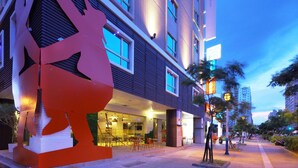 A modern building with a large abstract sculpture in front, a well-lit entrance, and a street with trees and parked cars.