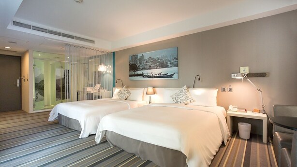A hotel room with two beds, a desk with a lamp, a small trash bin, and a painting on the wall.