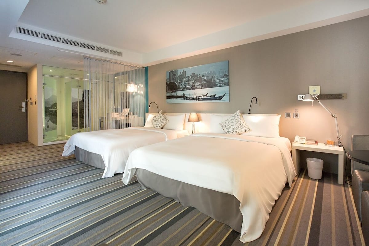 Een hotelkamer met twee bedden, een bureau met lamp, een kleine prullenbak en een schilderij aan de muur.