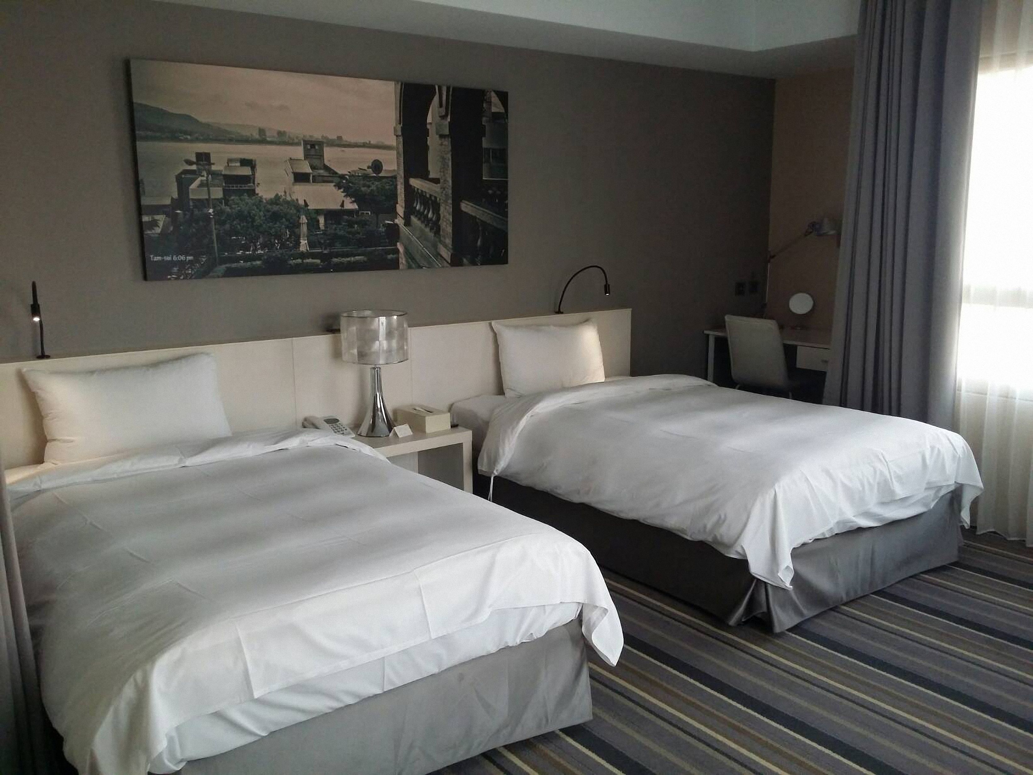 A hotel room with two beds, a desk, a lamp, and a painting on the wall.