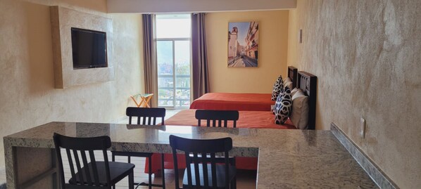 Habitación de hotel con cama, televisor montado en la pared, zona de comedor con mesa y sillas, y ventana con vista a edificios.
