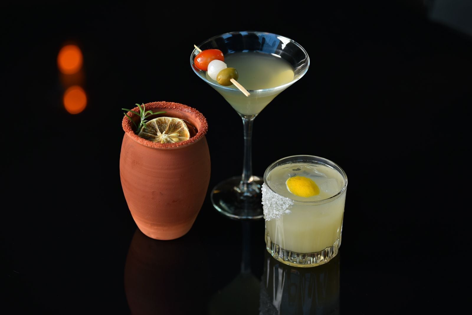 Three drinks: a martini, a cocktail with a lemon twist, and a shot glass with a lemon garnish.
