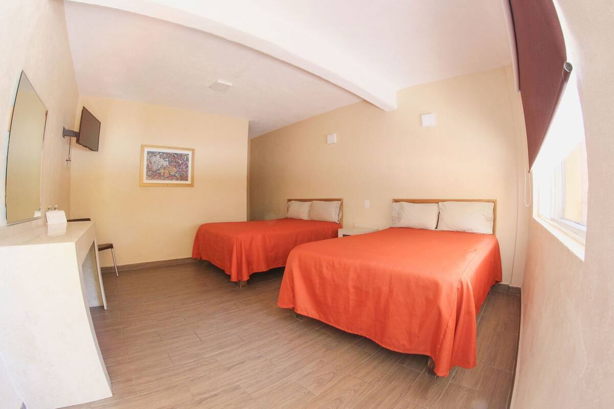 Una habitación con dos camas, un escritorio, un espejo, un cuadro en la pared y una ventana.